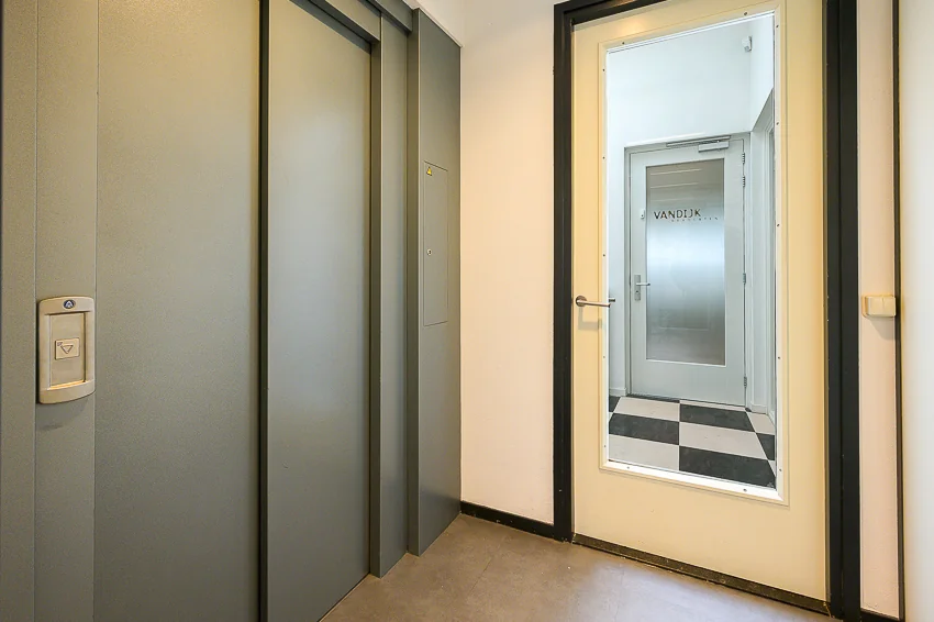 Hal met gesloten liftdeuren en een deur met raam dat uitzicht biedt op een gang met de naam 'VANDIJK' op een glazen deur.